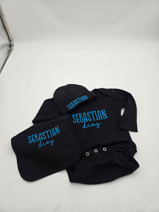 A baby clothing set laid flat on a white surface, including a black long-sleeve bodysuit, matching black bib, and black hat, each printed with the name 'SEBASTIAN' and the phrase 'diab' in bright blue lettering.
