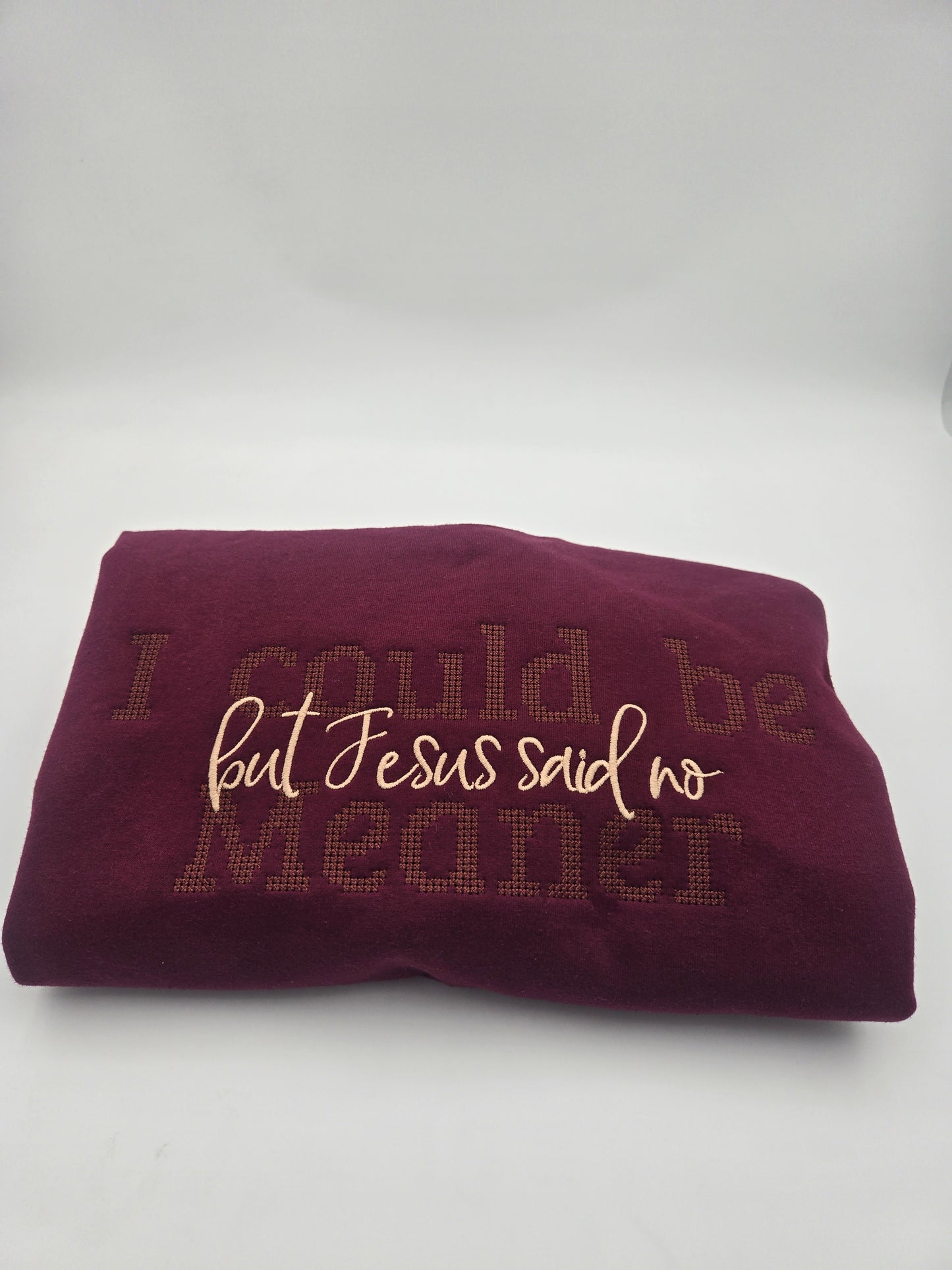 A folded burgundy sweater featuring the embroidered text 'I could be meaner but Jesus said no' in contrasting crean and brown tones, set against a plain light gray background.