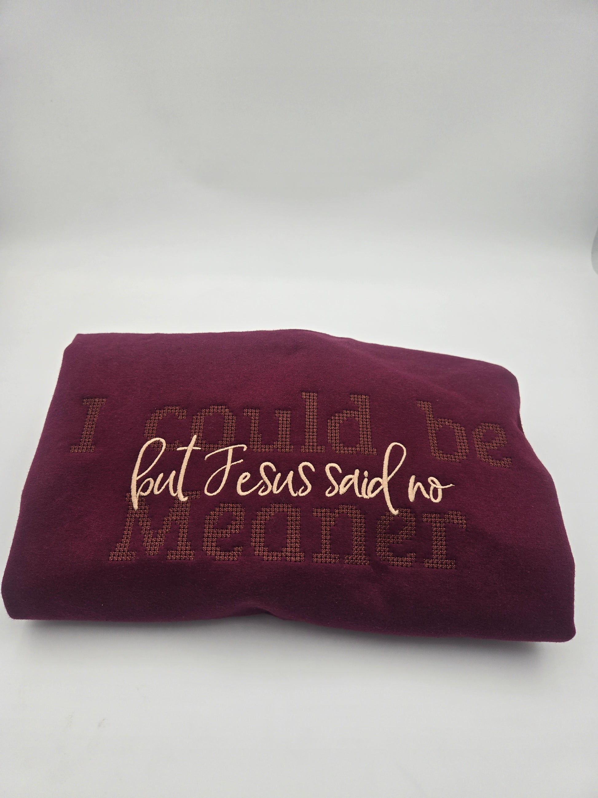 A folded burgundy sweater featuring the embroidered text 'I could be meaner but Jesus said no' in contrasting crean and brown tones, set against a plain light gray background.