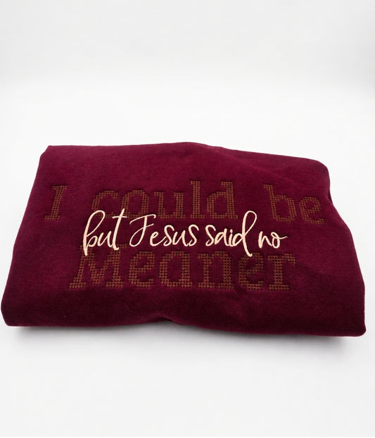 A folded burgundy fleece blanket featuring the embroidered text 'I could be meaner but Jesus said no' in cream and brown tones, set against a plain white background.
