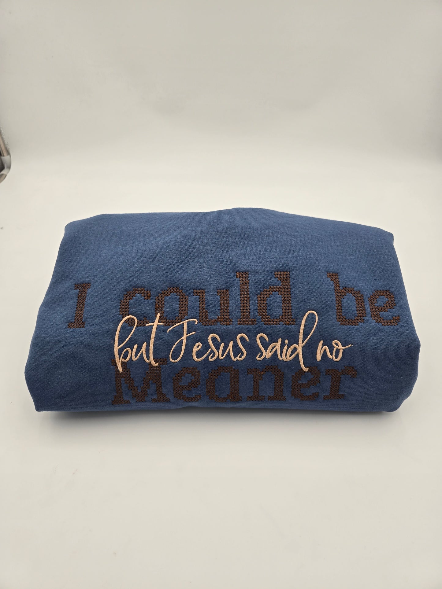 A folded indigo sweater with embroidered text reading 'I could be meaner but Jesus said no' in brown and light tan, shown on a plain light background.