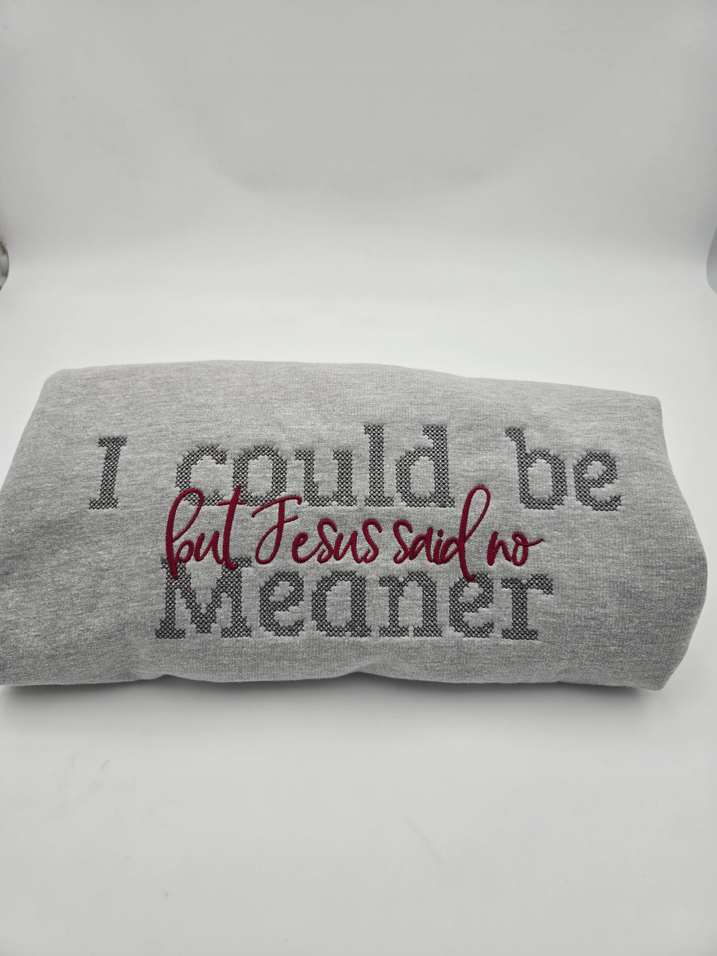 A folded gray heather sweatshirt with the text 'I could be Meaner but Jesus said no' in dark gray and burgundy embroidery, displayed on a white studio background.