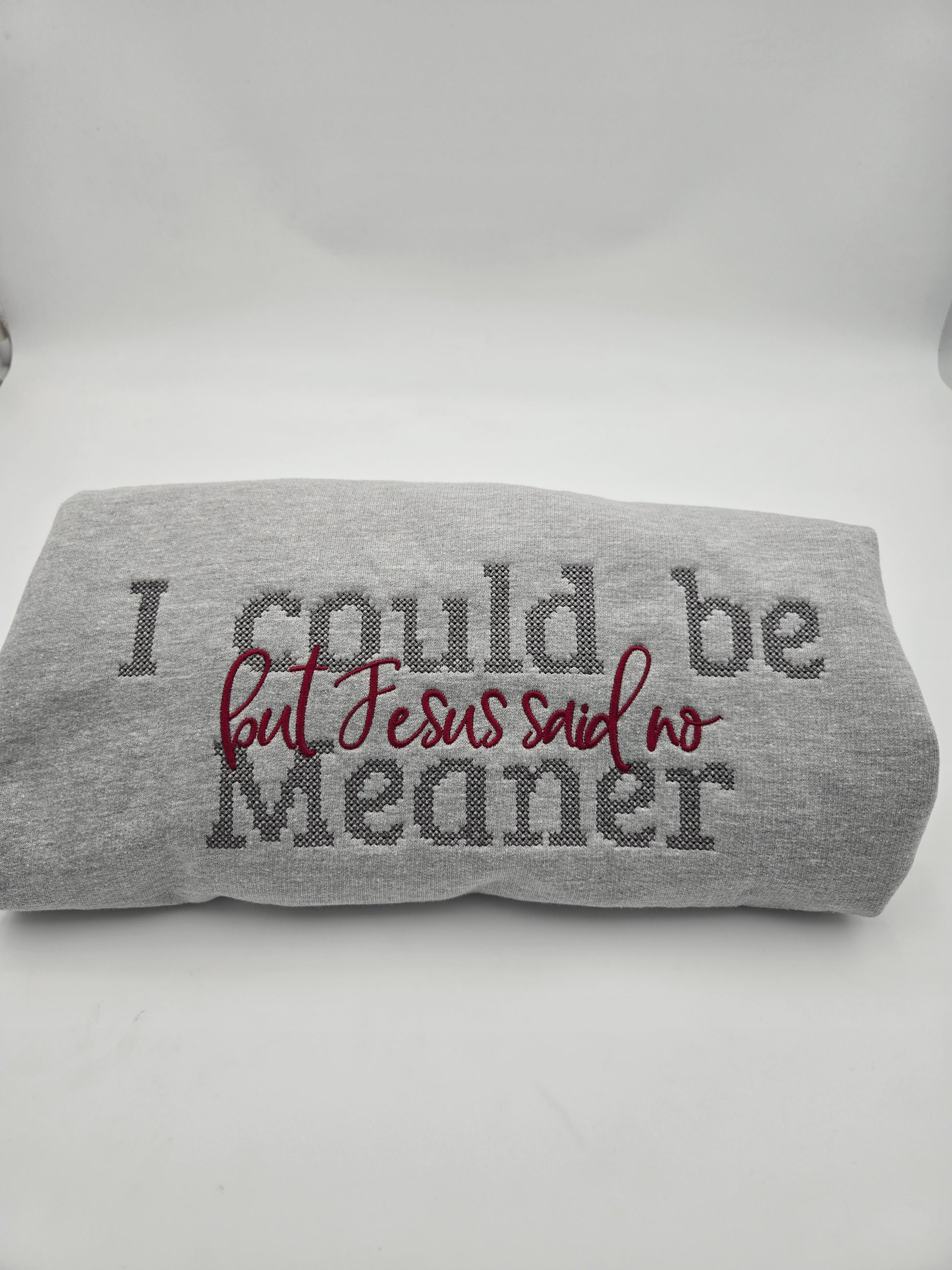 A folded gray heather sweatshirt with the text 'I could be Meaner but Jesus said no' in dark gray and burgundy embroidery, displayed on a white studio background.
