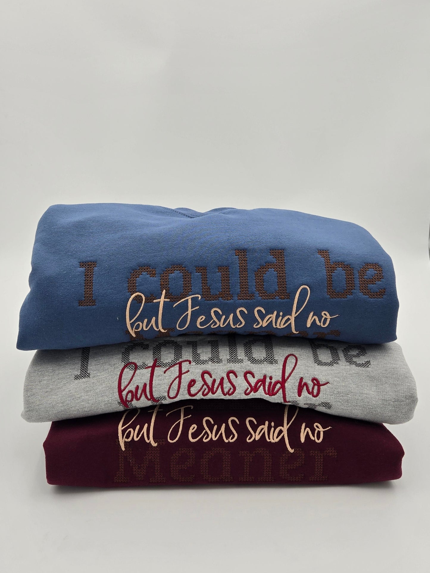 Three folded graphic t-shirts stacked on a white surface, featuring the text 'I could be but Jesus said no' in brown, gray, and burgundy lettering.