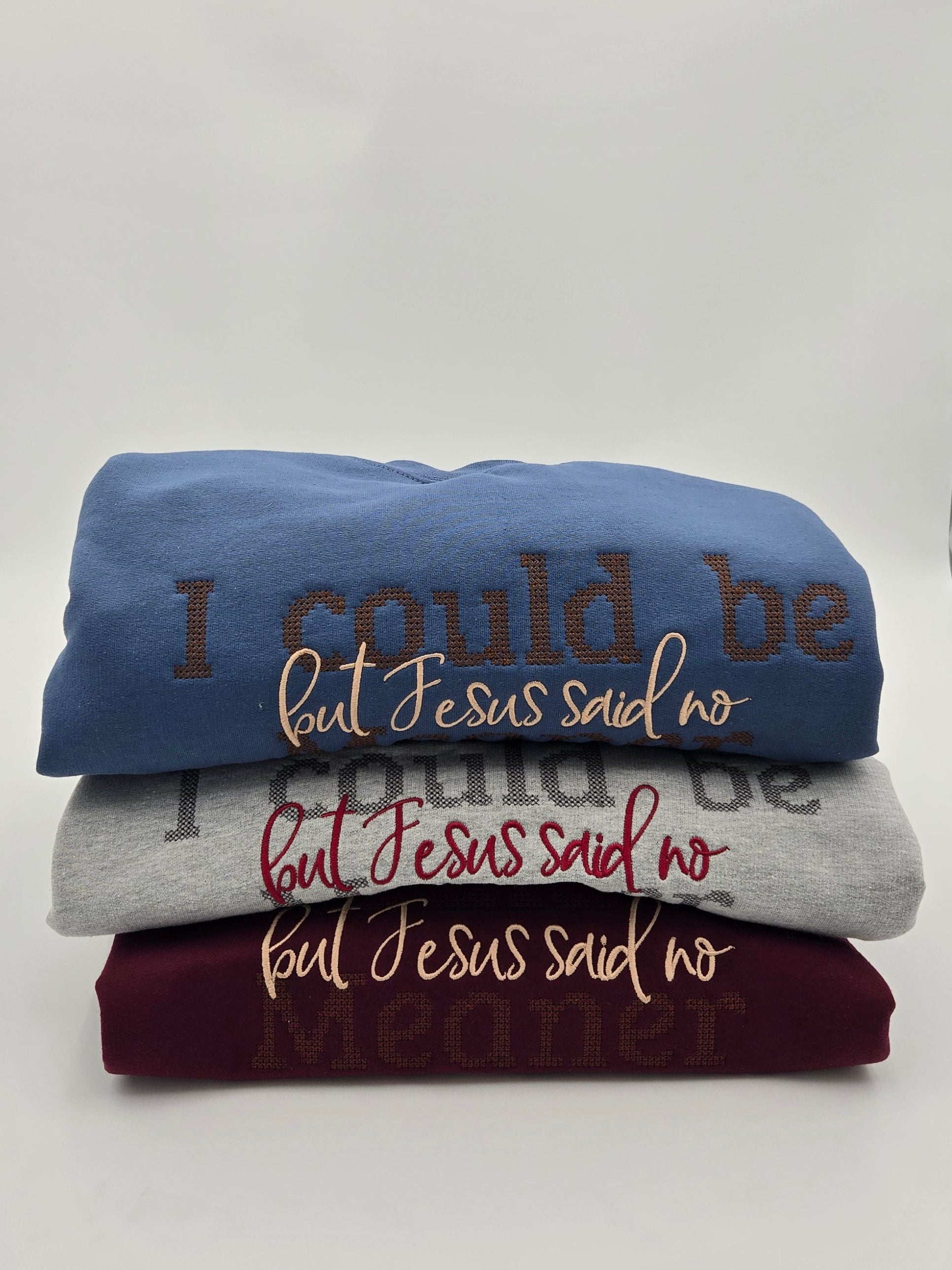 Three folded graphic t-shirts stacked on a white surface, featuring the text 'I could be but Jesus said no' in brown, gray, and burgundy lettering.