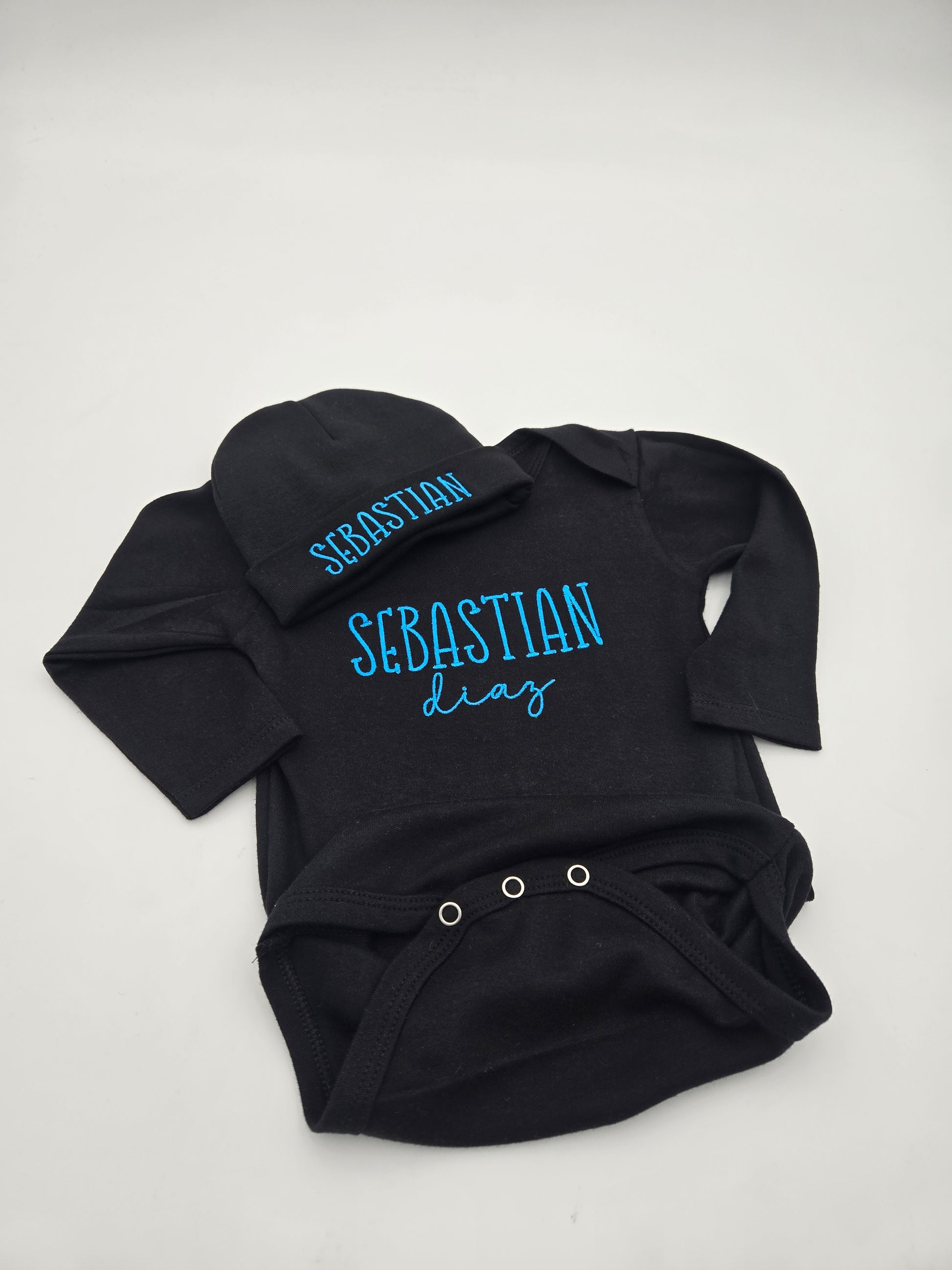 A black baby long-sleeve bodysuit with a matching beanie hat, both personalized with the name 'SEBASTIAN' in bright blue script. The bodysuit features 'SEBASTIAN' printed in blue on the chest and 'diaz' in a cursive font below it.